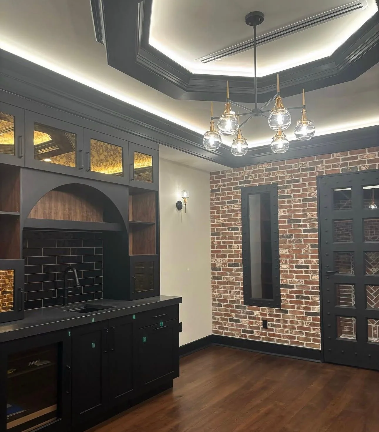 Speakeasy-style home bar with black cabinetry, brick wall, and cove-lit tray ceiling with invisible in-wall audio
