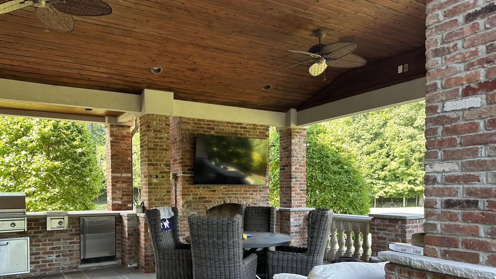 Covered patio with a TV mounted on a brick outdoor fireplace, ceiling fans, wicker seating, and a built-in outdoor kitchen with grill