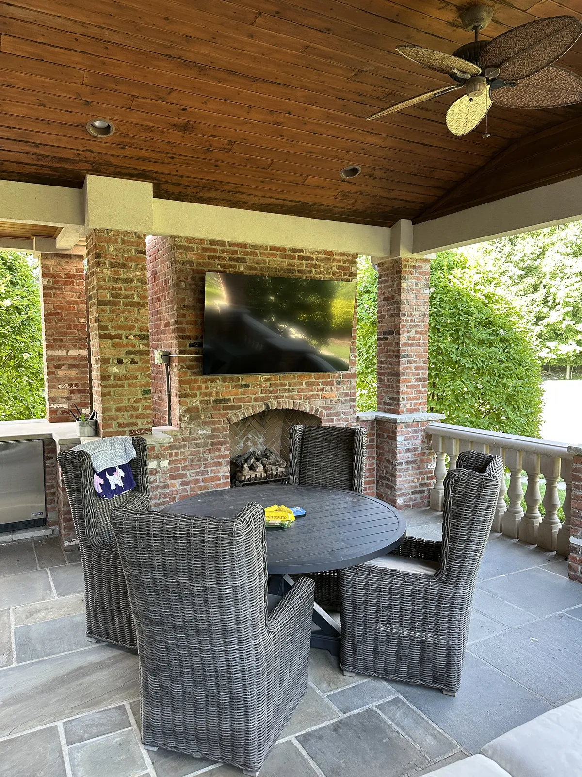 Covered patio with a weatherproof TV mounted on a brick outdoor fireplace, ceiling fans and wicker seating, in a NY/NJ home